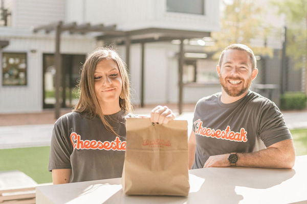 Guy and Girl wearing Philadelphia Cheesesteak shirts looking at a Deli Fresh Threads bag.